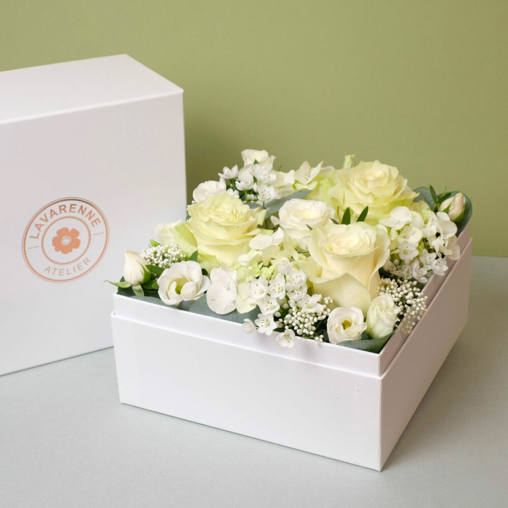 un coffret floral de l'Atelier Lavarenne, rempli de fleurs blanches et crème, dont des roses et des petites fleurs délicates, disposées soigneusement dans une boîte blanche. À gauche, une boîte blanche arbore le logo doré "LAVARENNE ATELIER" avec une fleur au centre. Le tout est placé sur un fond vert pastel, donnant une sensation de fraîcheur et d'élégance.