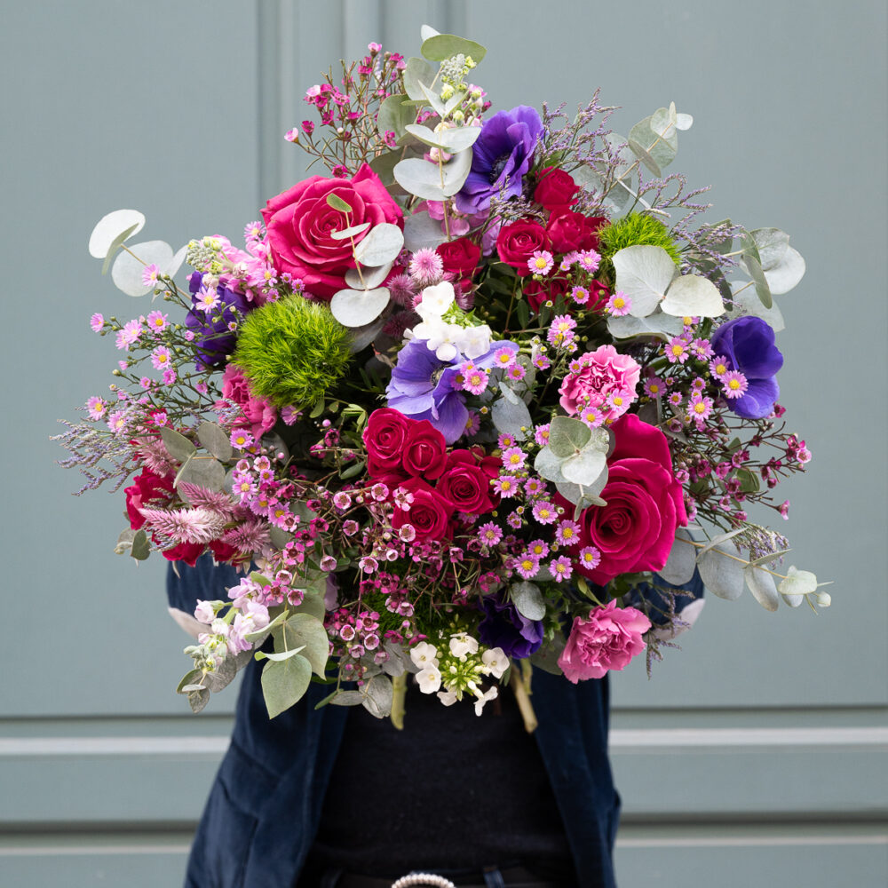 Une personne portant un grand bouquet aux tons roses, rouges et violets, capturé de face devant une porte vert clair.