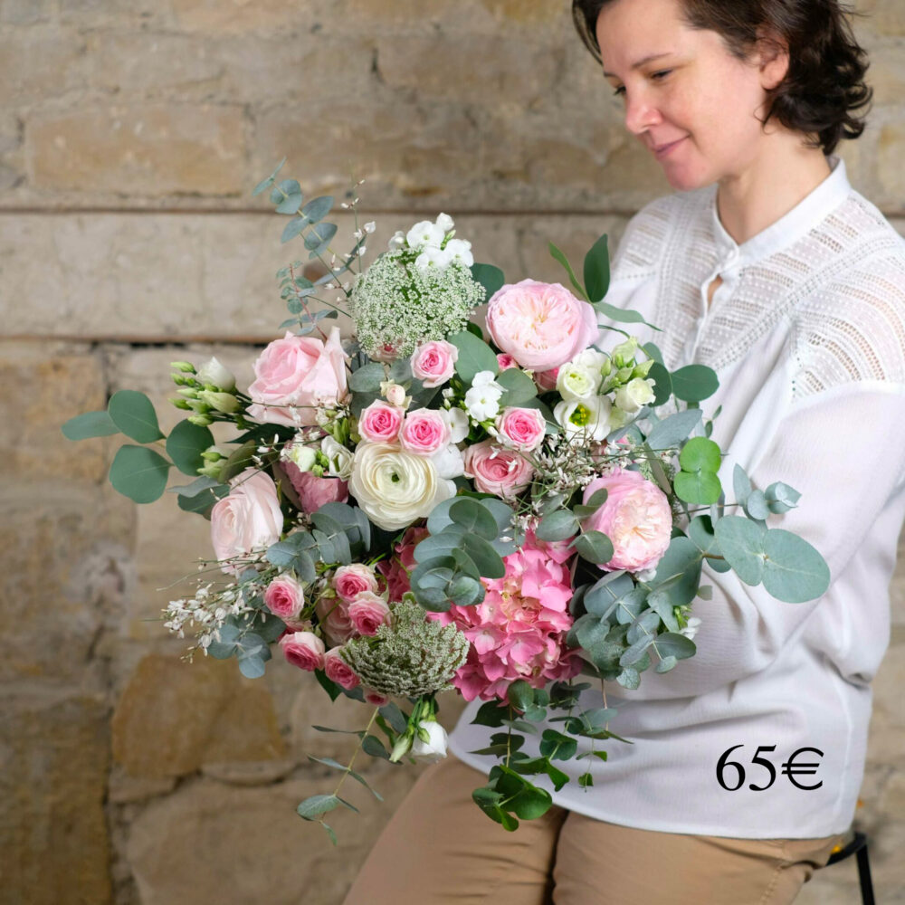 une femme en blouse blanche tenant un grand bouquet luxuriant de fleurs aux couleurs pastel, comprenant des roses roses et blanches, des renoncules, des hortensias et des feuilles d'eucalyptus. Le bouquet, délicat et romantique, est affiché au prix de "65€" dans le coin inférieur droit. En arrière-plan, un mur de pierre rustique ajoute une touche naturelle et sereine à la scène.