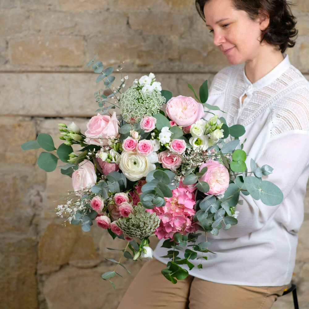 une femme en blouse blanche tenant un grand bouquet de fleurs aux tons pastels, incluant des roses roses et blanches, des renoncules, des hortensias roses et des feuilles d'eucalyptus. Le fond est un mur de pierre rustique, ajoutant une ambiance naturelle et douce à la scène. Le bouquet donne une impression de tendresse et d'élégance.