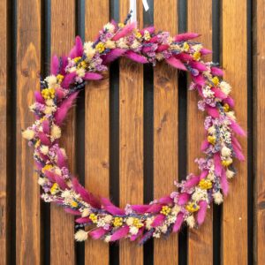 une couronne de fleurs séchées suspendue sur un mur en bois. La couronne est composée de plumes roses vives, de petites fleurs violettes et jaunes, et d'épis de blé doré, créant un contraste vibrant avec le fond en bois naturel. L'agencement des couleurs et des textures donne à la couronne une apparence festive et chaleureuse.