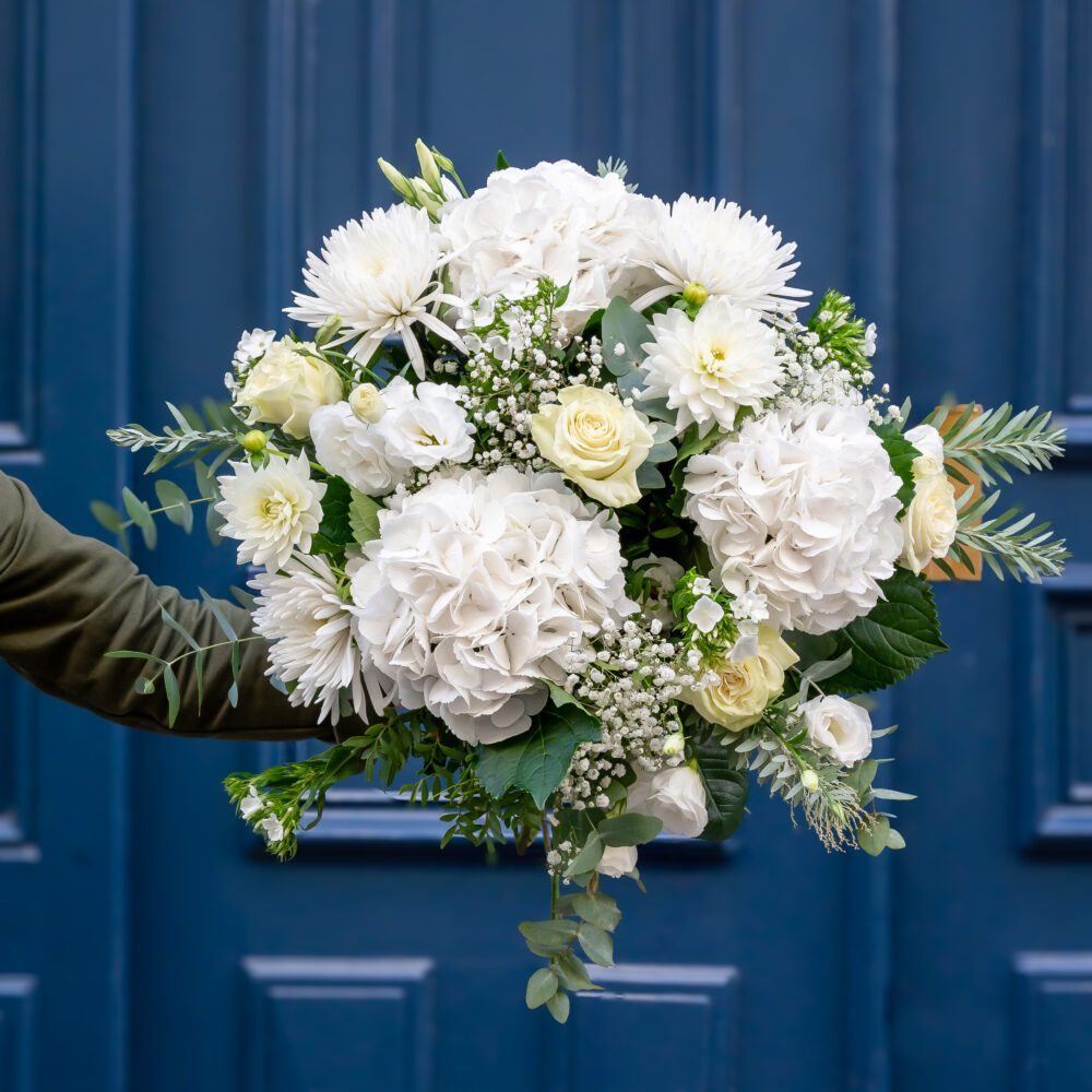 un grand bouquet de fleurs blanches comprenant des roses, des hortensias et d'autres fleurs délicates, tenu par une personne vêtue d'une manche verte. Le bouquet est volumineux et raffiné, avec des feuillages verts ajoutant du contraste. En arrière-plan, une porte bleu foncé donne une touche élégante et urbaine à la présentation florale.