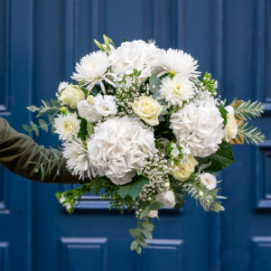 un grand bouquet de fleurs blanches comprenant des roses, des hortensias et d'autres fleurs délicates, tenu par une personne vêtue d'une manche verte. Le bouquet est volumineux et raffiné, avec des feuillages verts ajoutant du contraste. En arrière-plan, une porte bleu foncé donne une touche élégante et urbaine à la présentation florale.