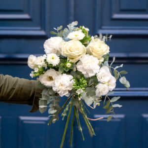 un bouquet de fleurs blanches comprenant des roses, des œillets et d'autres petites fleurs délicates, tenu par une personne portant une manche verte. Le bouquet est entouré de feuillage vert, ajoutant une texture naturelle à l'ensemble. En arrière-plan, une porte bleu foncé accentue le contraste avec les fleurs, offrant une présentation élégante et sobre.