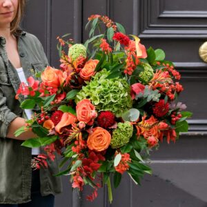 L'image montre une femme tenant un grand bouquet de fleurs aux tons orange, vert et rouge devant une porte noire. Le bouquet est composé de roses orange vif, d'hortensias verts, de baies rouges et de feuillage vert, avec des touches de fleurs rouges et des éléments de texture comme des baies et des feuilles colorées.