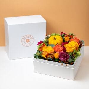 un coffret floral de l'Atelier Lavarenne, composé de fleurs aux couleurs automnales, notamment des roses jaunes, des œillets oranges, des fleurs rouges et une mini-citrouille. Le coffret est placé devant une boîte blanche portant le logo "LAVARENNE ATELIER", sur un fond orange clair.