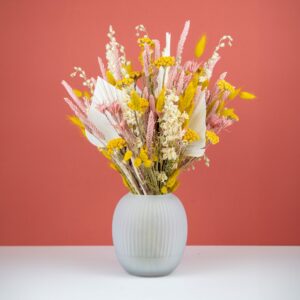 un bouquet de fleurs séchées dans un vase en verre dépoli, disposé sur une surface blanche. Le bouquet est composé de tiges jaunes, roses et blanches, avec des accents de feuilles de palmier et de petits épis, apportant un mélange de textures et de couleurs douces. Le fond corail accentue l'éclat des couleurs naturelles du bouquet.