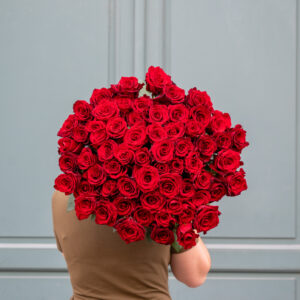 une personne tenant un grand bouquet de roses rouges devant une porte bleu-vert. Le bouquet est composé de nombreuses roses rouges parfaitement arrangées, créant un effet volumineux et spectaculaire.