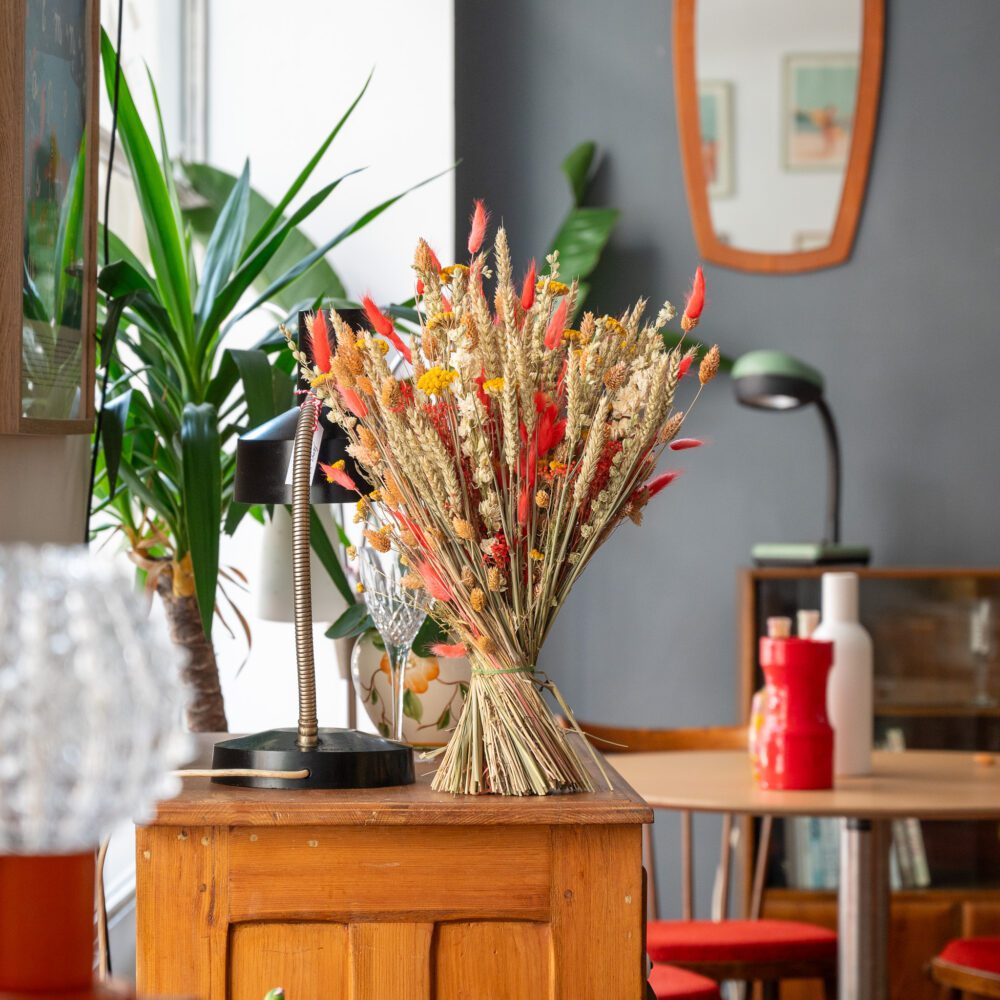 un bouquet de fleurs séchées disposé sur un meuble en bois dans un intérieur lumineux. Le bouquet est composé de blé doré, de petites fleurs jaunes et de plumes rouges, apportant une touche de couleur naturelle à la pièce. En arrière-plan, on voit des plantes vertes, une lampe de bureau noire, un miroir mural, ainsi qu'une table avec des objets décoratifs, créant une ambiance chaleureuse et accueillante.