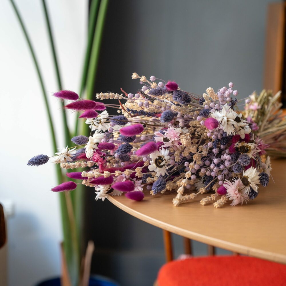 un bouquet de fleurs séchées posé sur une table en bois clair. Le bouquet est composé de lavande, de marguerites blanches, de petites fleurs violettes, et de plumes roses vives. En arrière-plan, on aperçoit une plante verte et une chaise avec un coussin orange, créant une ambiance chaleureuse et naturelle.