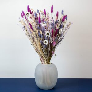 un bouquet de fleurs séchées dans un vase en verre dépoli. Le bouquet est composé de lavande, de marguerites blanches, de petites fleurs violettes, et de plumes roses vives. Le vase est posé sur une surface bleu marine, créant un contraste élégant avec le fond blanc minimaliste.