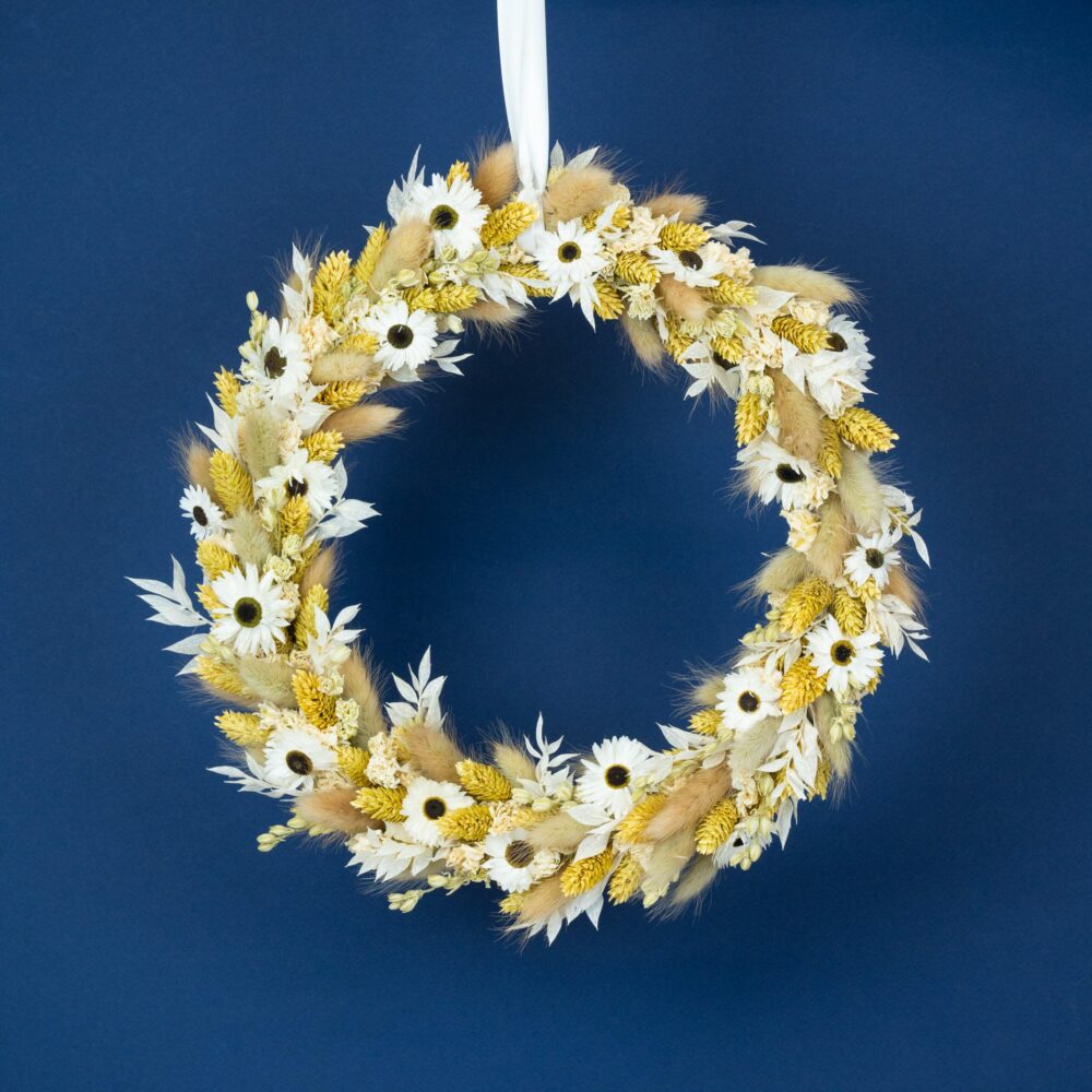 une couronne de fleurs séchées suspendue par un ruban blanc. La couronne est composée de marguerites blanches, d'épis de blé doré, de feuilles blanches et de brins d'herbe douce. Le fond bleu foncé met en valeur les tons naturels et doux de la couronne.