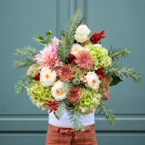 Personne tenant un grand bouquet de fleurs composé de roses blanches, d'hortensias verts, de chrysanthèmes roses et saumon, avec des touches de feuillage vert et rouge, devant une porte bleu-vert