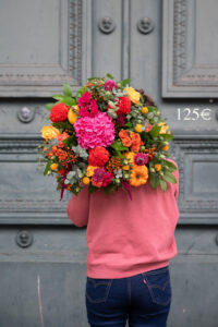 une personne portant un grand bouquet de fleurs aux couleurs vives (rouge, rose, orange et jaune) comprenant des hortensias, des roses, des dahlias, et une petite citrouille. La personne porte un haut rose et se tient devant une porte gris-vert. Le prix "125€" est inscrit en haut à droite.