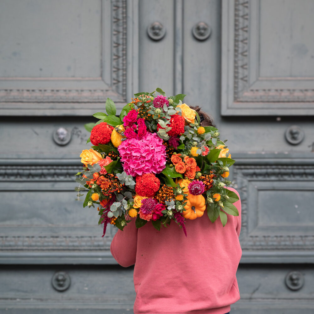 une personne de dos, vêtue d'un haut rose, portant un grand bouquet coloré de fleurs d'automne. Le bouquet est composé de roses, d'hortensias, de dahlias et de divers feuillages, dans des tons vifs de rouge, rose, orange et jaune. On peut également voir une mini-citrouille intégrée à la composition. L'arrière-plan est une porte ancienne gris-vert, ajoutant un contraste élégant aux couleurs éclatantes du bouquet.