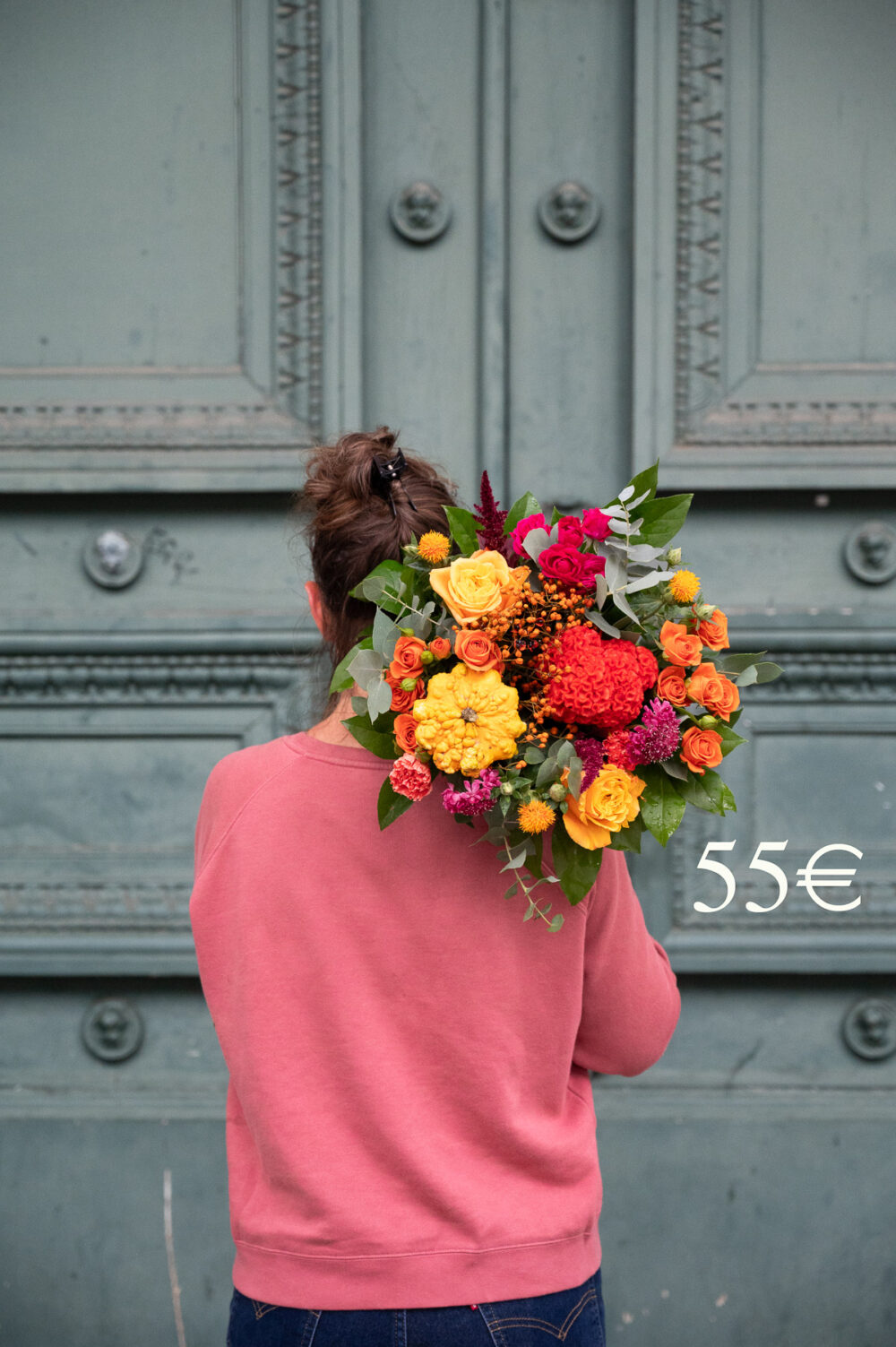 une personne portant un bouquet de fleurs plus petit, également aux tons rouges, oranges et jaunes, composé de roses, de fleurs de saison et de verdure. La personne est également vêtue d'un haut rose et se tient devant une porte de couleur similaire. Le prix affiché est "55€".