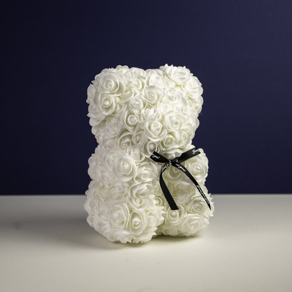 un ours en forme de peluche entièrement composé de roses artificielles blanches. L'ours est décoré d'un ruban noir noué autour de son cou, avec une inscription délicate en écriture cursive. Le fond est d'un bleu marine profond, contrastant avec l'éclat des roses blanches.