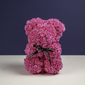 un ours en forme de sculpture, composé de petites roses roses vives. Un ruban noir avec l'inscription "Just for you" en blanc est noué autour de son cou. Le fond violet foncé met en valeur les détails floraux et la couleur vibrante de l'ours.