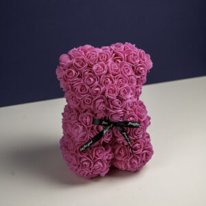 un ours en forme de sculpture, composé de petites roses roses vives. Un ruban noir avec l'inscription "Just for you" en blanc est noué autour de son cou. Le fond violet foncé met en valeur les détails floraux et la couleur vibrante de l'ours.