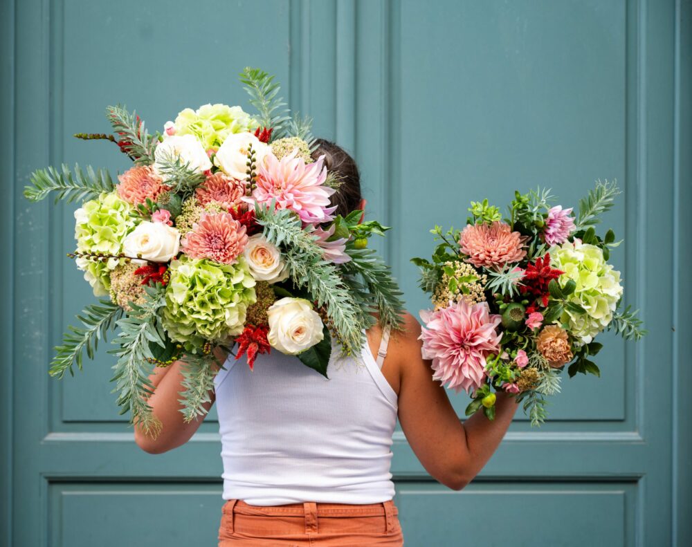 Personne tenant deux grands bouquets de fleurs variées, incluant des hortensias verts, des dahlias roses, des roses blanches et d'autres fleurs colorées, devant une porte de couleur bleu-vert