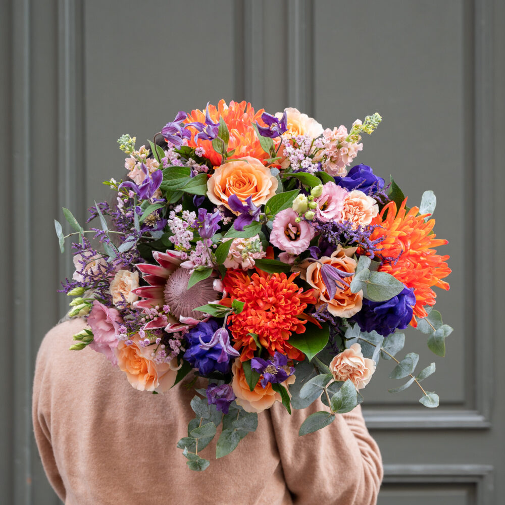 Bouquet du mois de novembre avec des fleurs éclatantes en tons orange, violet et rose, comprenant des roses, des proteas, et des chrysanthèmes, tenu par une personne en pull beige devant une porte verte.