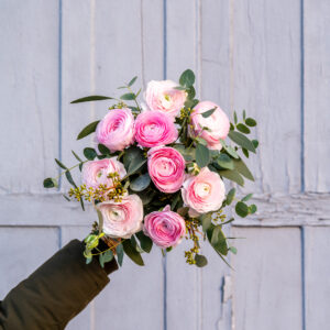 Un bouquet de renoncules roses et blanches accompagné de feuillage vert, tenu à bout de bras devant une porte en bois bleu vieilli.