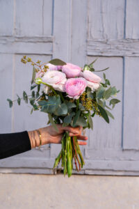 Une main tenant un bouquet de renoncules roses et blanches, leurs tiges longues et feuillues apparaissant clairement devant un décor de porte bleu patiné.